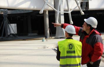 Two men in hard hats standing together while one points at something in the distance.