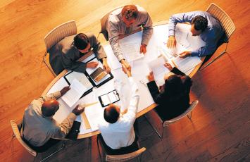Group of business people sitting at a large table reviewing paperwork with two of the people shaking hands over the table
