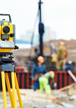 A theodolite in focus with people in the distance working and viewing the area.