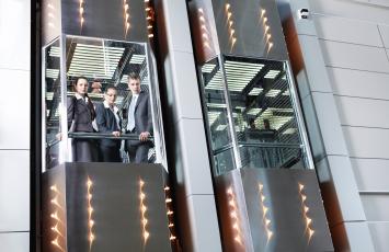An elevator with three business people peering below through the glass as the use the elevator